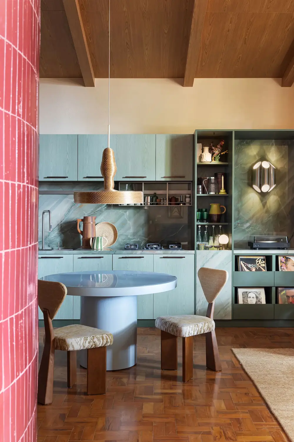 Contemporary Brazilian kitchen with sage green cabinetry, a marble backsplash, a round pedestal dining table, sculptural wooden pendant light, parquet flooring, and exposed timber ceiling beams.