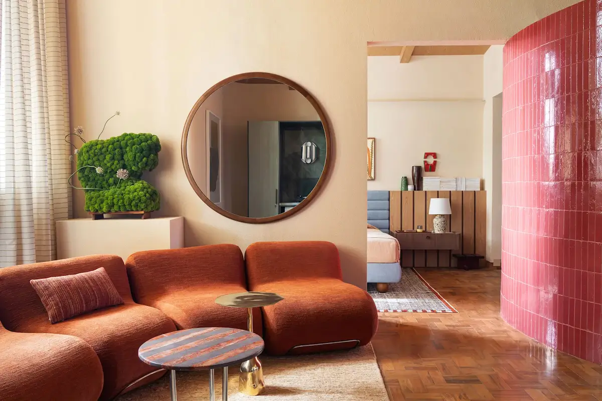 Warm living room in a small Brazilian apartment with a curved terracotta sofa, a round wall mirror, a glossy pink tiled column, parquet flooring, and a view into the bedroom.
