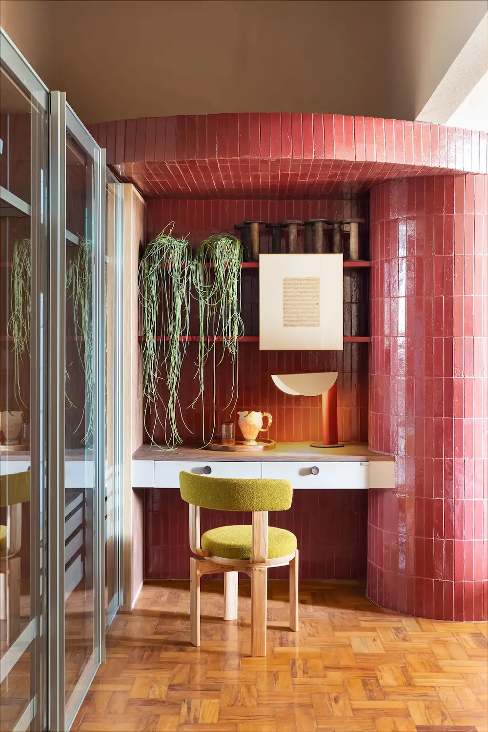 Compact home office nook in a Brazilian apartment featuring glossy terracotta tiled walls, a built-in desk, an olive green upholstered chair, open shelving, hanging plants, and warm parquet flooring.