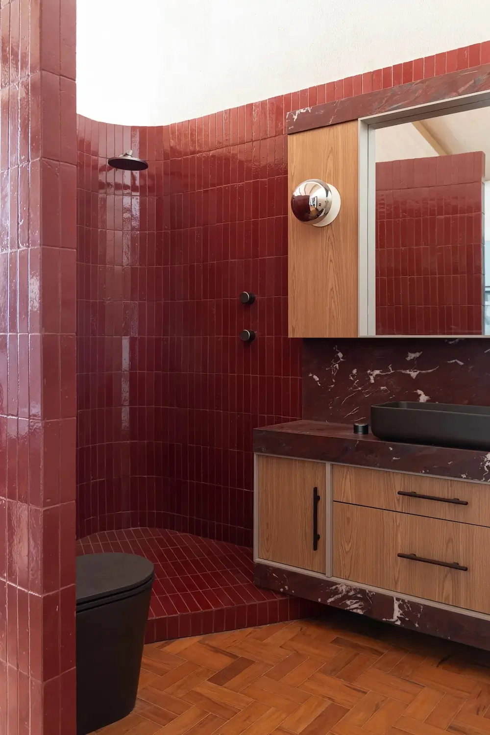 Modern bathroom in a Brazilian apartment with glossy burgundy tiled walls, a walk-in shower, a floating oak vanity, a dark stone countertop, and matte black fixtures.