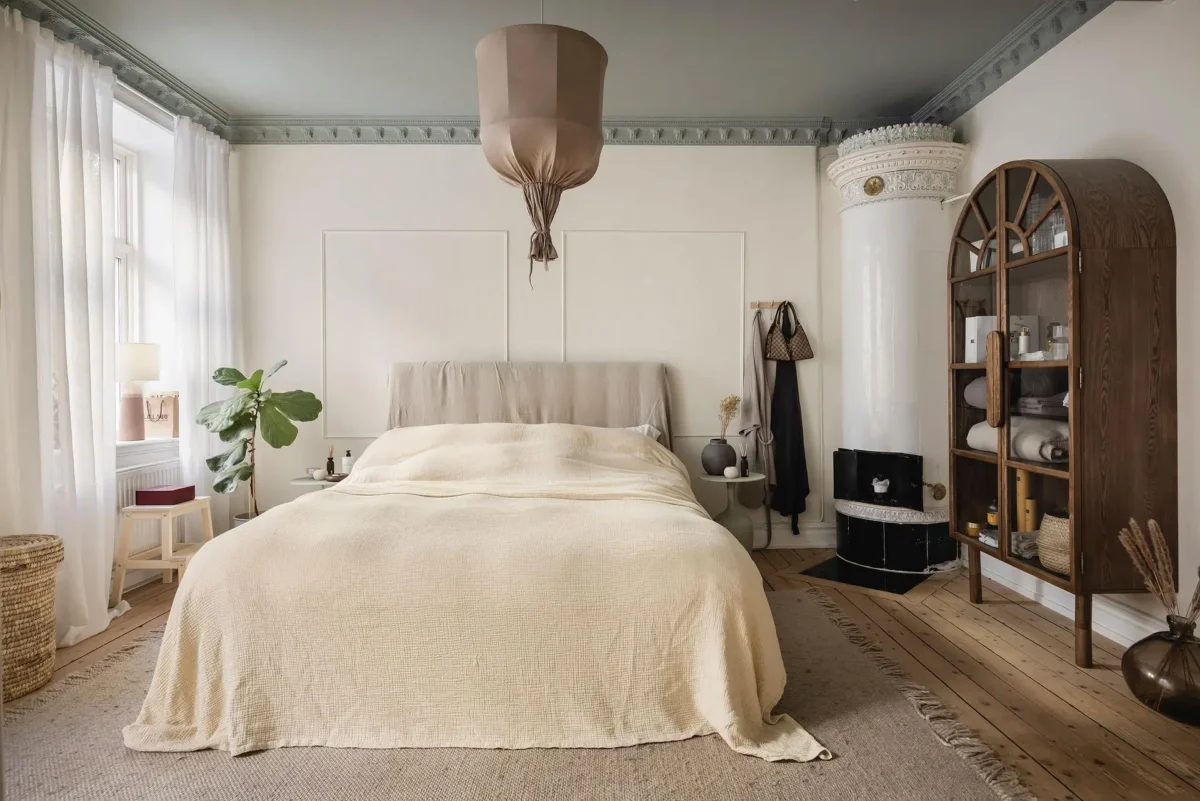 Alt text: Bright neutral bedroom in a 1905 Swedish apartment with cream bedding, original tiled stove, decorative moldings, wooden floors and an arched cabinet.