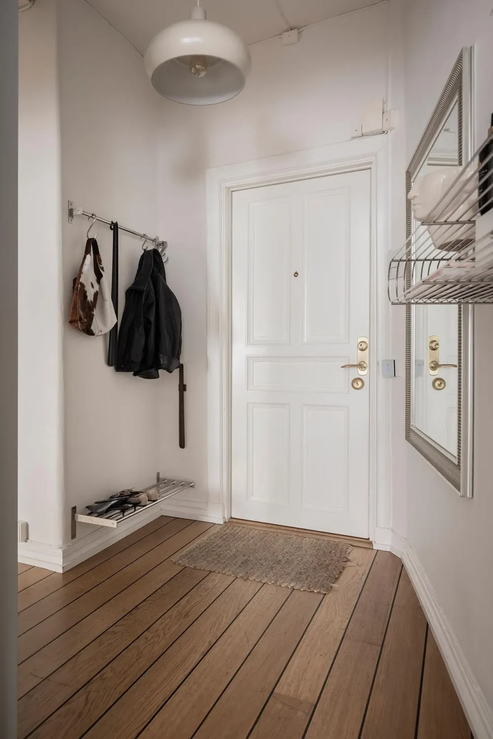 Minimal Scandinavian entryway with white paneled door, warm wooden floors, simple wall hooks, metal shoe rack and a large framed mirror.