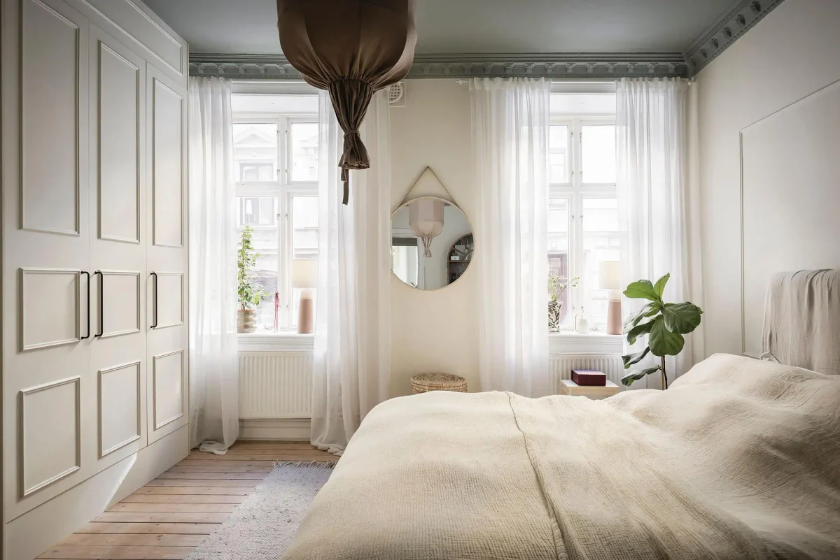 Light-filled Swedish bedroom with cream bedding, paneled wardrobes, sheer white curtains, pale wooden floors and soft gray-green ceiling moldings.