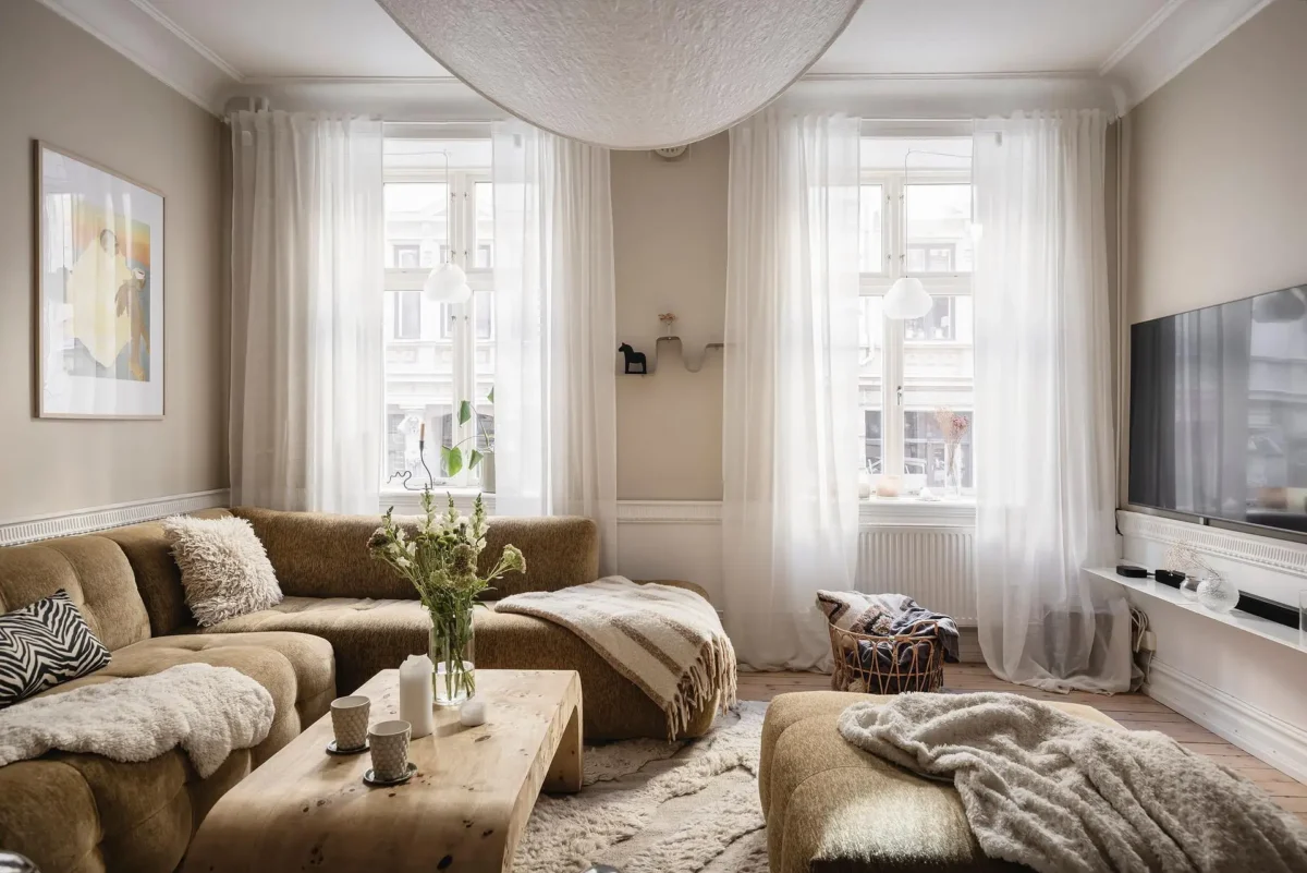 Cozy neutral Scandinavian living room with a curved beige sofa, oversized ottoman, wooden coffee table, layered textiles and sheer white curtains.
