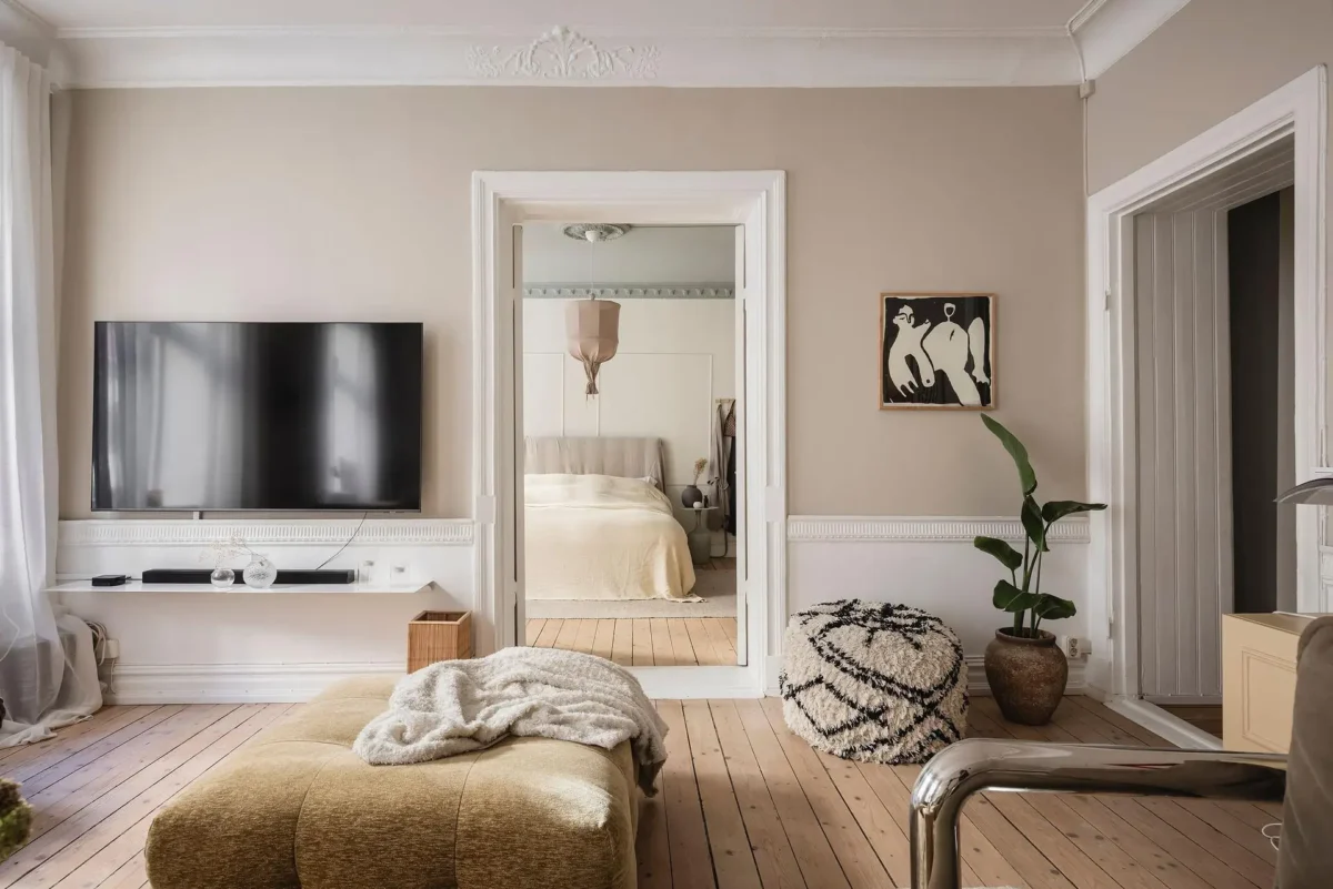 Neutral Scandinavian living room with warm beige walls, wooden floors, textured ottoman, Moroccan-style pouf and a view into the adjoining bedroom.
