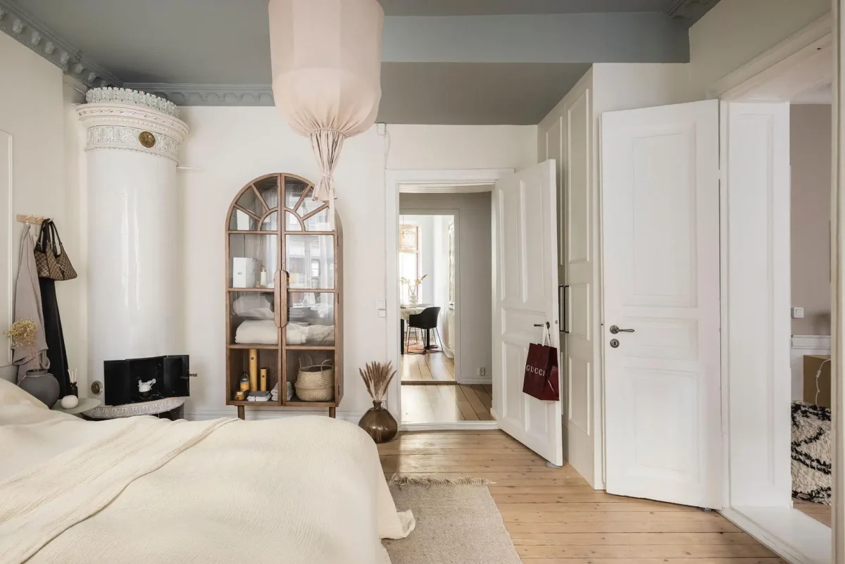 Neutral Swedish bedroom with an ornate tiled stove, arched wooden cabinet, cream bedding, original wooden floors and decorative gray-green ceiling moldings.