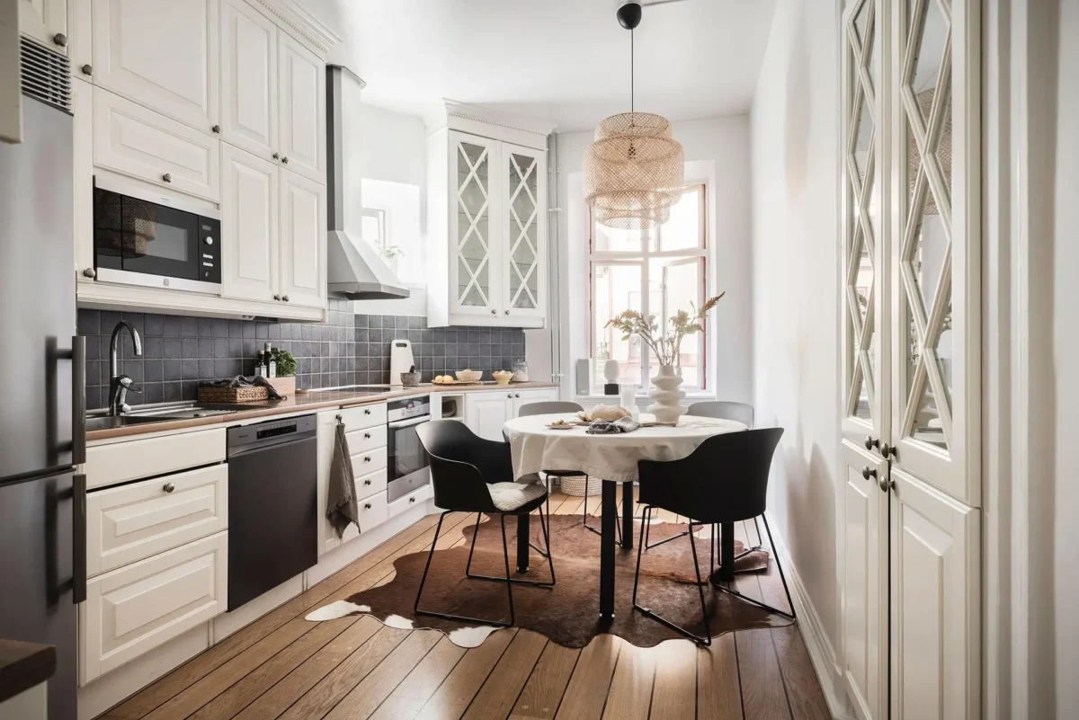 Bright Scandinavian kitchen with white paneled cabinets, dark tile backsplash, wooden countertops, round dining table and a woven pendant light.