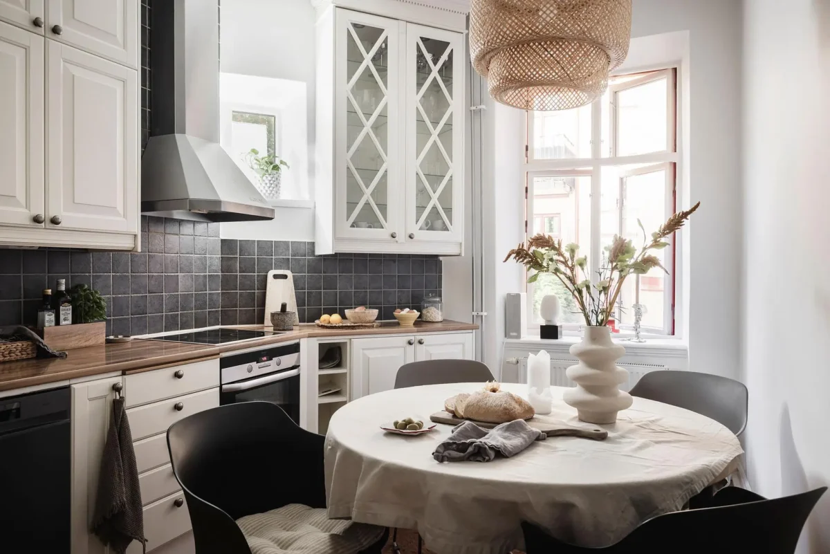 Scandinavian kitchen with white paneled cabinets, dark square tile backsplash, wooden countertops and a round dining table beneath a woven pendant light.