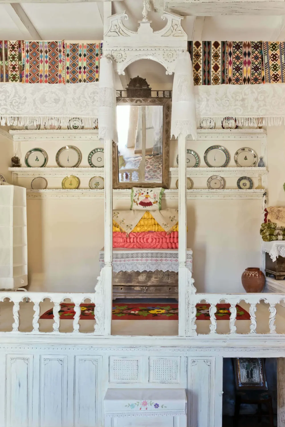 Traditional Karpathian raised sleeping platform with white-painted carved woodwork, colorful woven textiles, embroidered linens and antique ceramic plates.