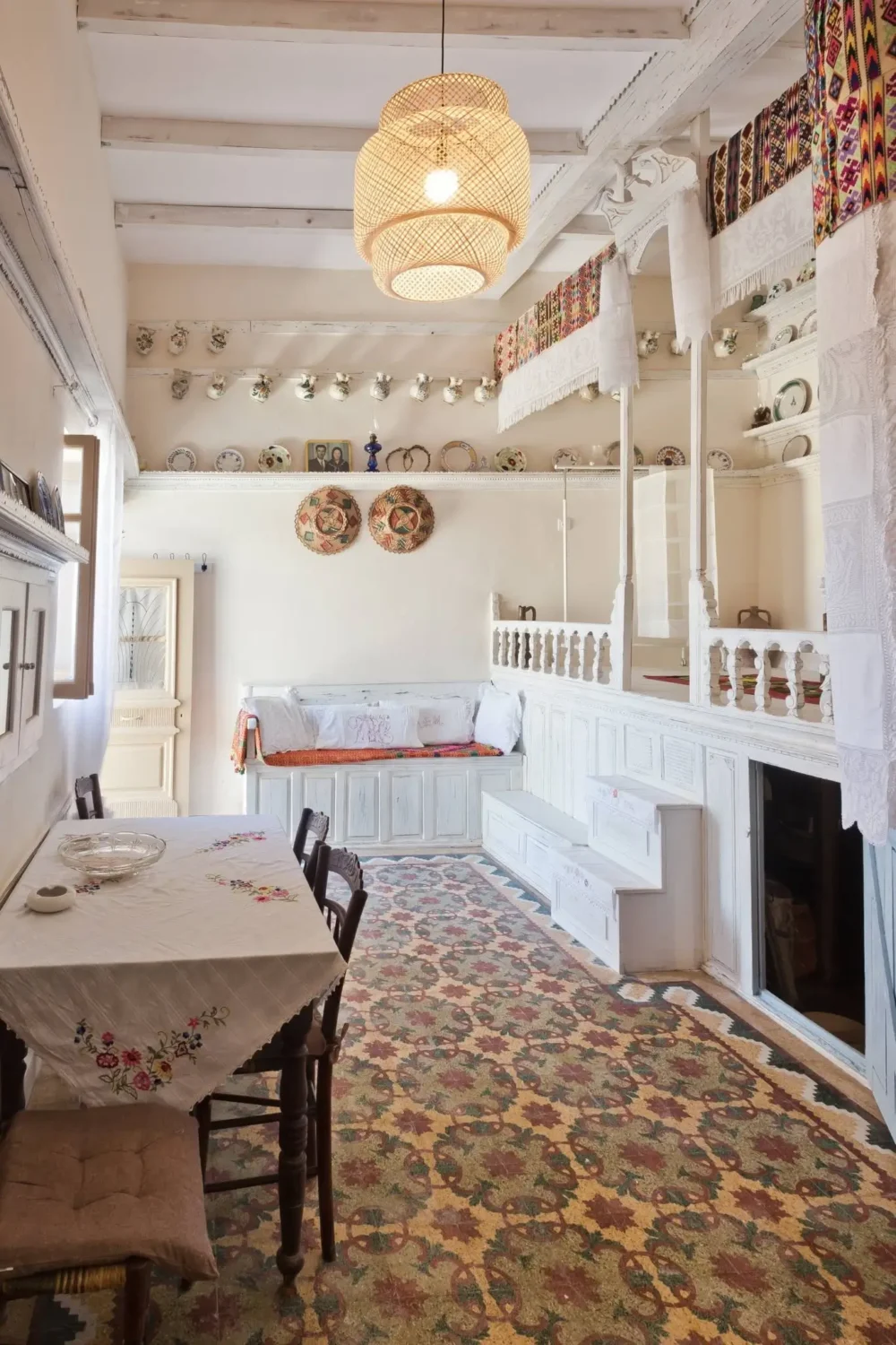Traditional Karpathian interior in Menetes, Greece, with white-painted carved woodwork, raised platform, built-in seating, embroidered textiles, antique ceramics and a colorful patterned mosaic floor.