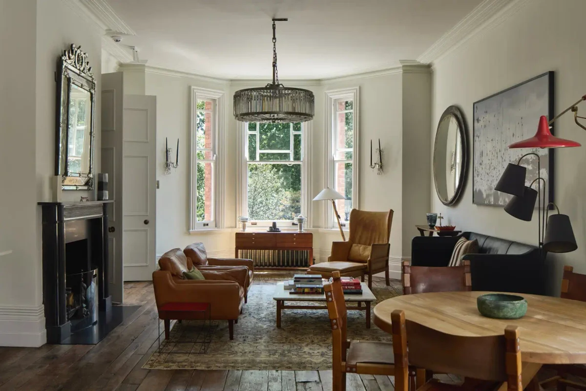 Elegant historic living room with original ceiling moldings, bay windows, dark wooden floors, a black fireplace, and modern furniture mixed with vintage pieces.