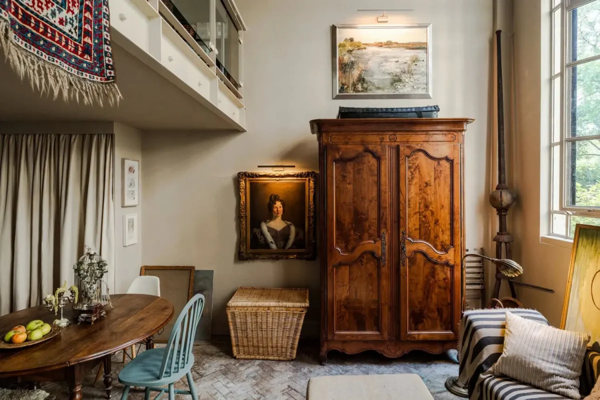 Historic London apartment with an antique wooden armoire, vintage portrait, mismatched dining chairs, worn parquet flooring, and a modern mezzanine.