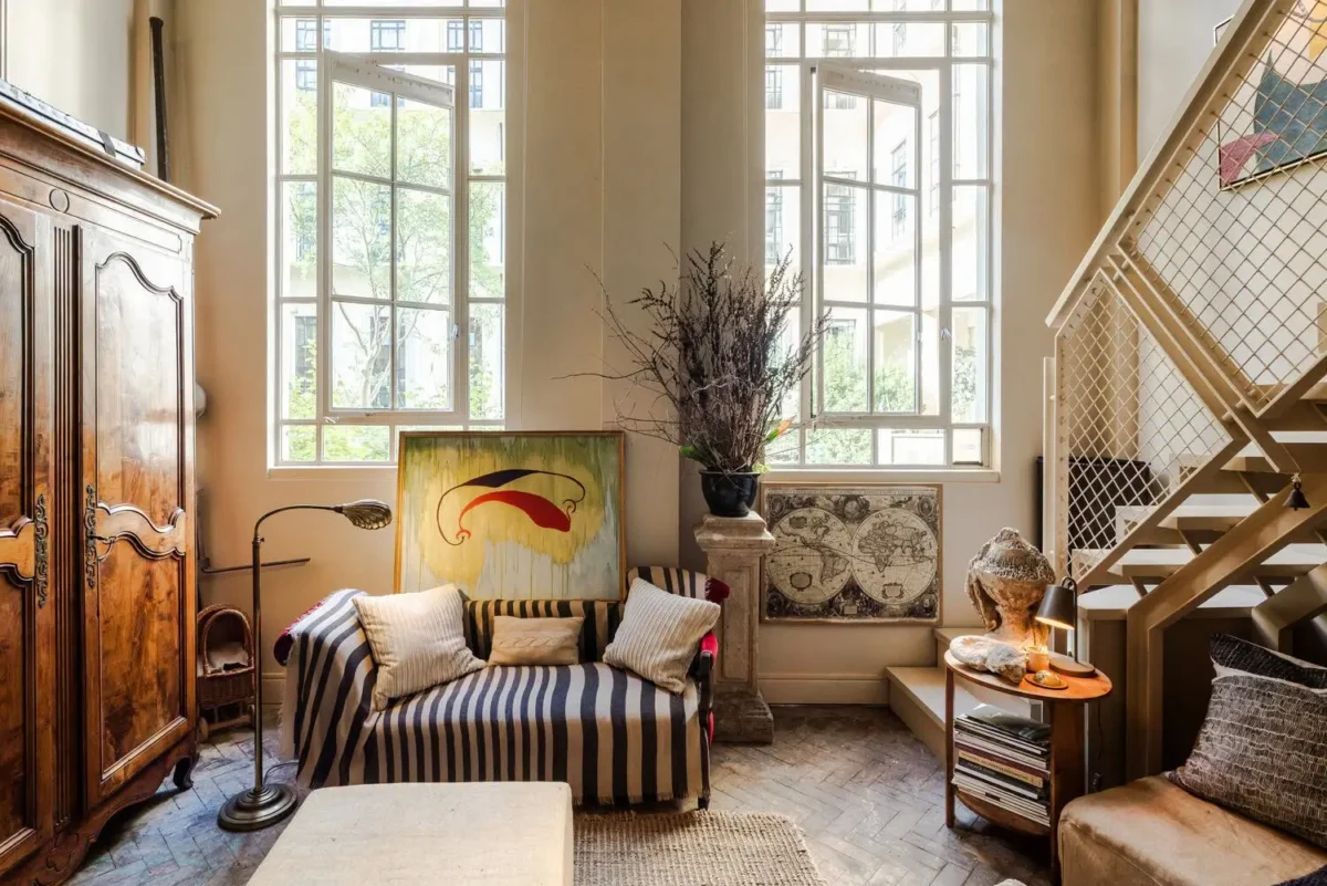 Historic London apartment with tall windows, aged parquet flooring, an antique wooden armoire, striped sofa, modern artwork, and a contemporary metal staircase.